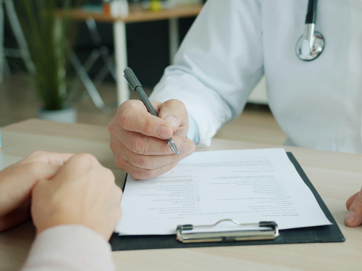 A physician signing a medical form on a clipboard across the desk from a patient